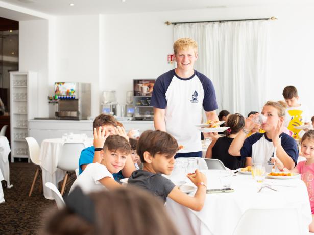 Un gruppo di bambini mangia in un ristorante con un cameriere sorridente.