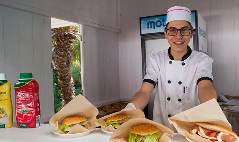 Lächelnder Koch serviert Burger und Hotdog an einem Außenkiosk.