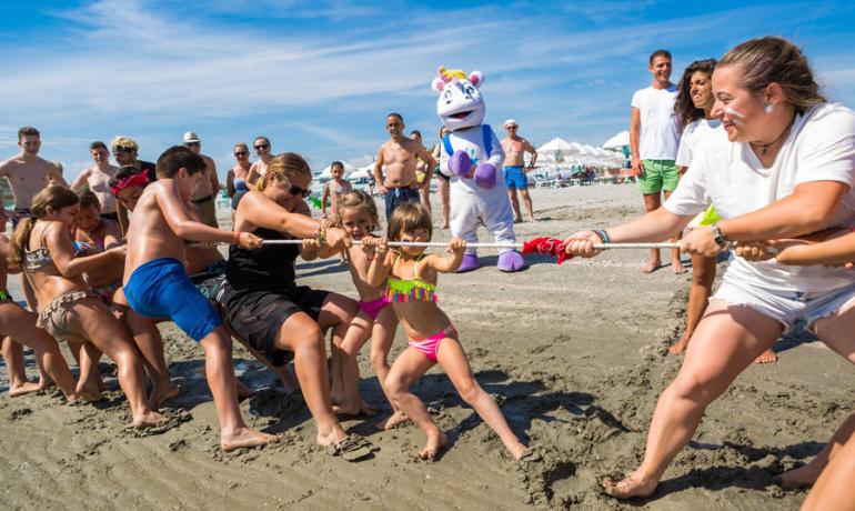 Gara di tiro alla fune sulla spiaggia con persone e una mascotte.