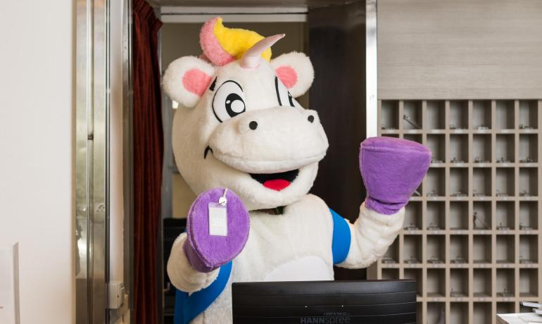 Un unicorno mascotte con guanti viola alla reception di un hotel.