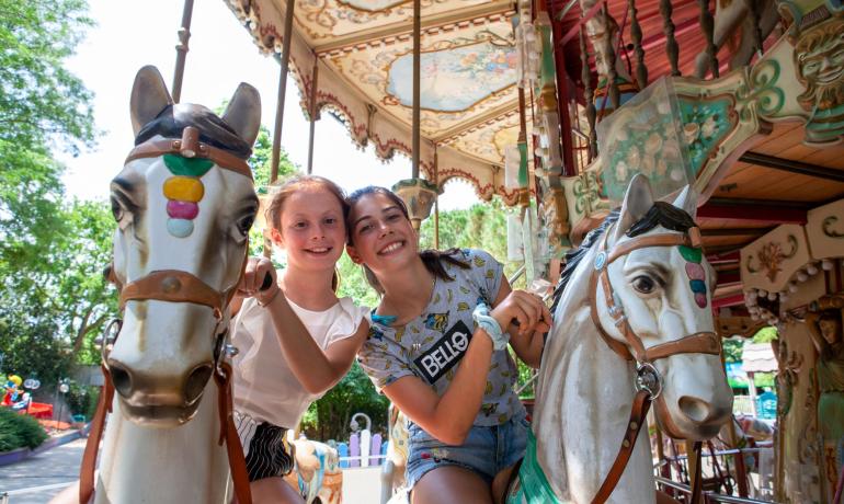 Two smiling girls on a colorful carousel, surrounded by a joyful atmosphere.