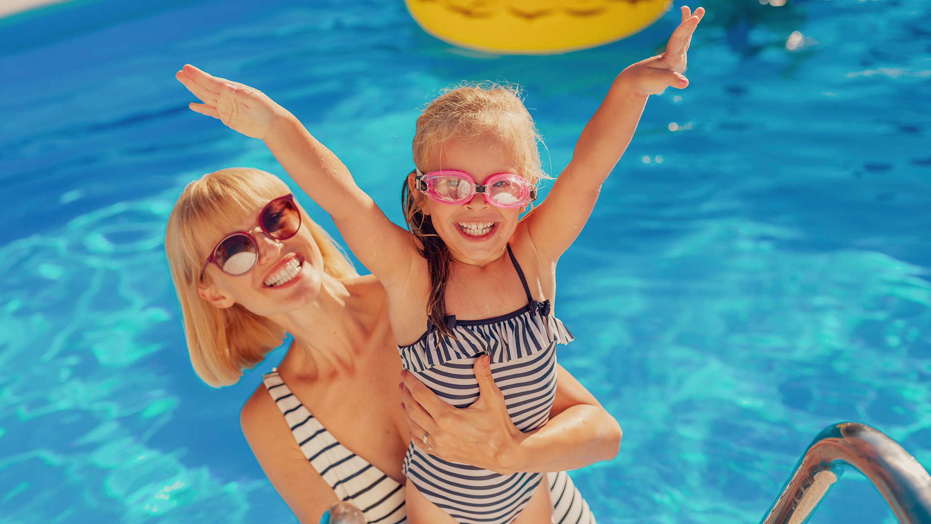 Bambino e uomo in piscina con braccioli, ombrelloni colorati sullo sfondo.