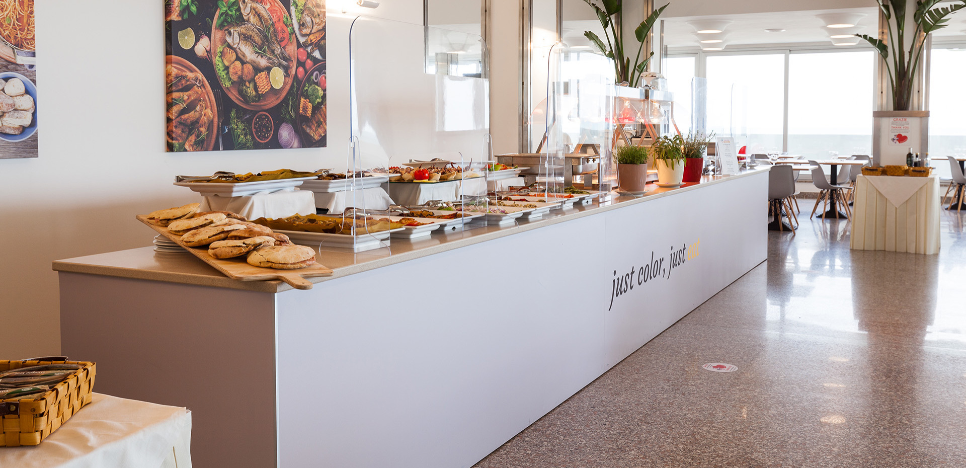 Buffet con pizza e verdure, chef sullo sfondo in cucina luminosa.