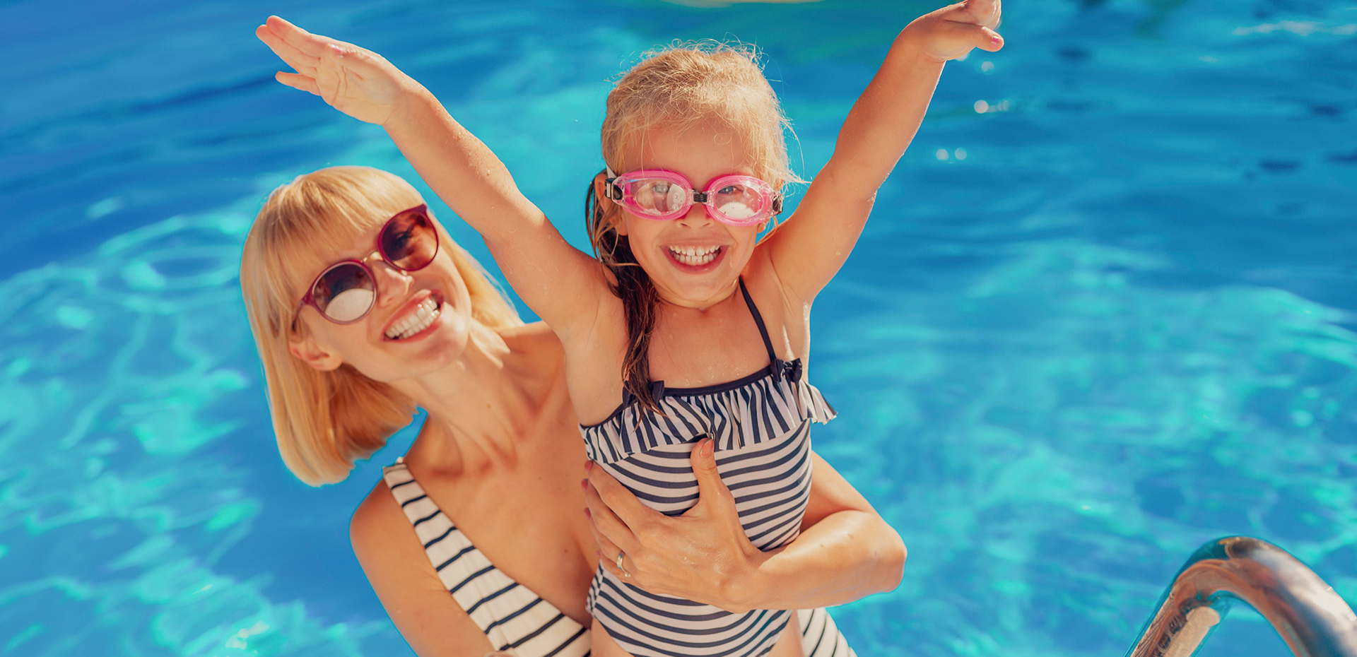 Bambino e uomo in piscina con braccioli, ombrelloni colorati sullo sfondo.