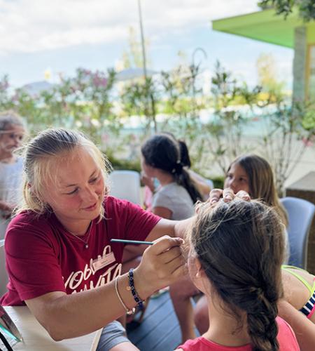 Bambino seduto al tavolo con colori, accompagnato da un adulto sorridente.