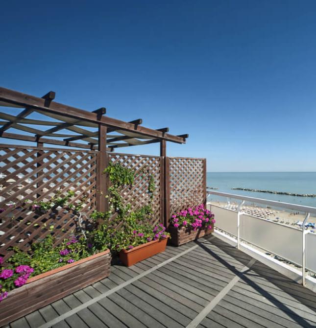 Terrazza fiorita con vista mare, sole splendente e spiaggia affollata.